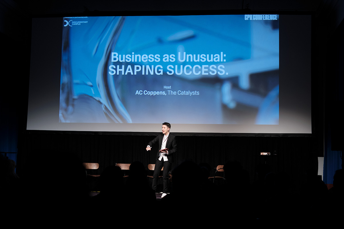AC Coppens, wearing a Black suit, speaks to the audience at the 2022 CPH: Conference while standing in front of a digital screen. Courtesy of CPH: Conference.