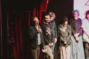 Photograph of a group of people standing on stage. One white man is holding a microphone and smiling at the audience.
