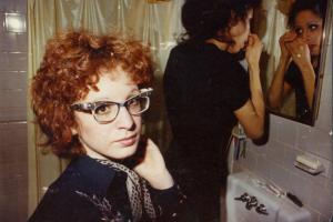 A woman wearing black adjusts her eye makeup in the mirror, while a red-headed woman wearing glasses looks at the camera in the foreground. From ‘All the Beauty and the Bloodshed.’ Photo courtesy of Neon. 