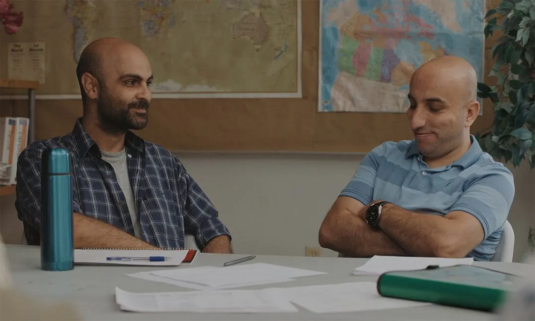 Image featuring two men sitting next to each other at a table in a school room.