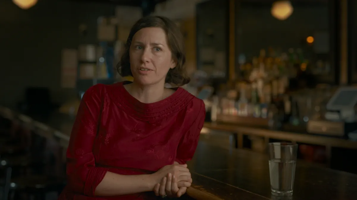 A woman in a red blouse sits leaning against a bar countertop, speaking to an interviewer behind the camera.