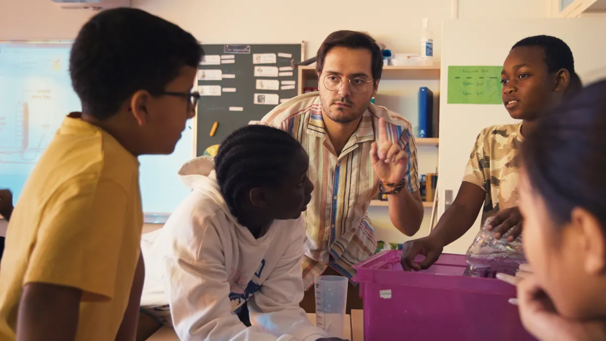 In a bright classroom, a teacher holds up his finger in front of a table of students.