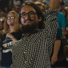 Film still from 'Your Friend, Memphis,' depicting a smiling young white man in a checkered shirt, raising his left arm, in a crowded stadium.