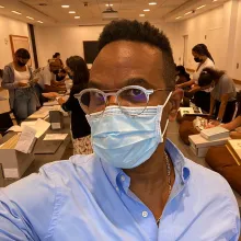 Thomas Allen Harris is a Black male filmmaker, seen here wearing a blue shirt, glasses, and a surgical mask. He is taking a selfie, and his students are visible in the background. Courtesy of Thomas Allen Harris.